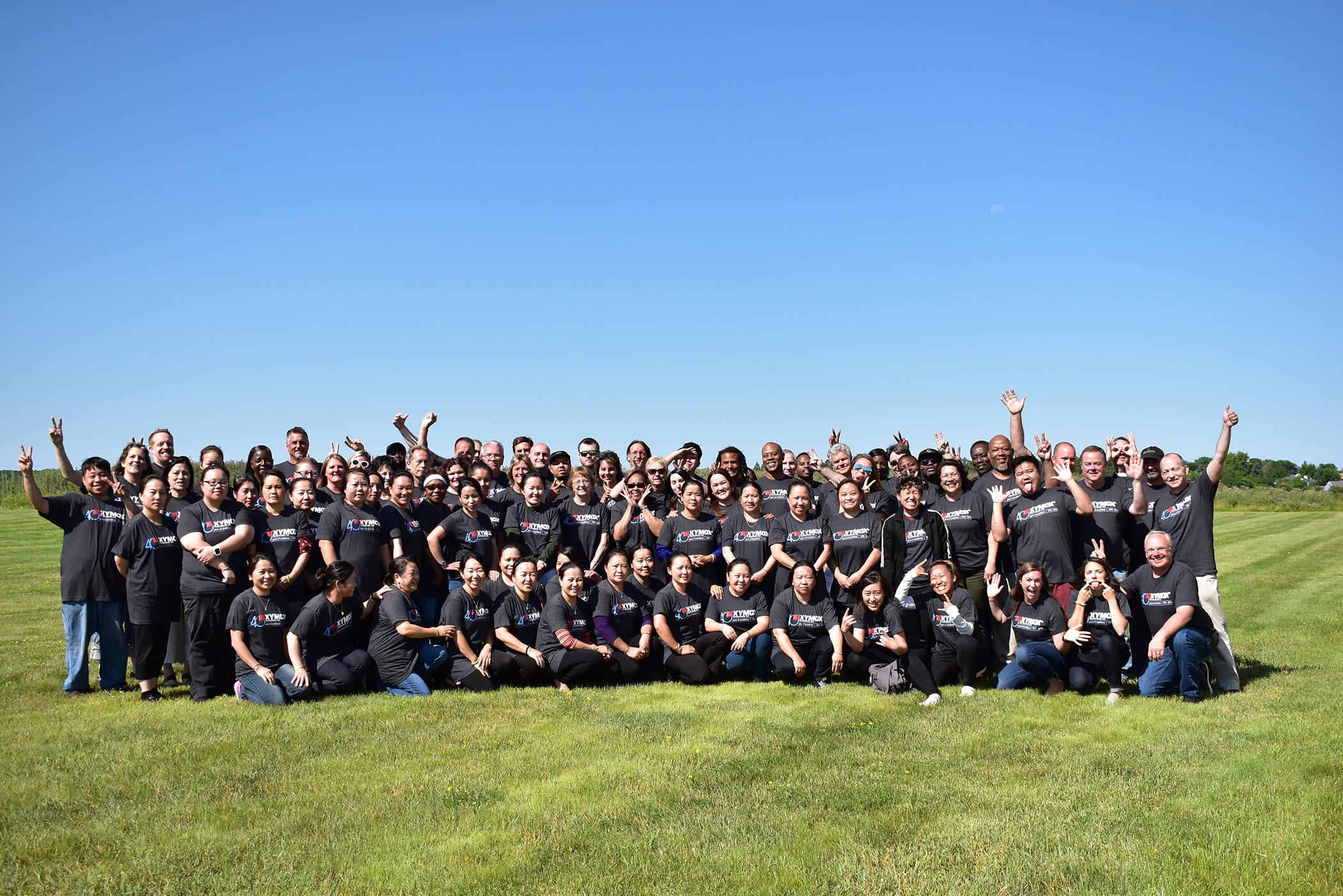 100+ Xymox employees, all wearing a gray company shirt and posing for a fun group picture