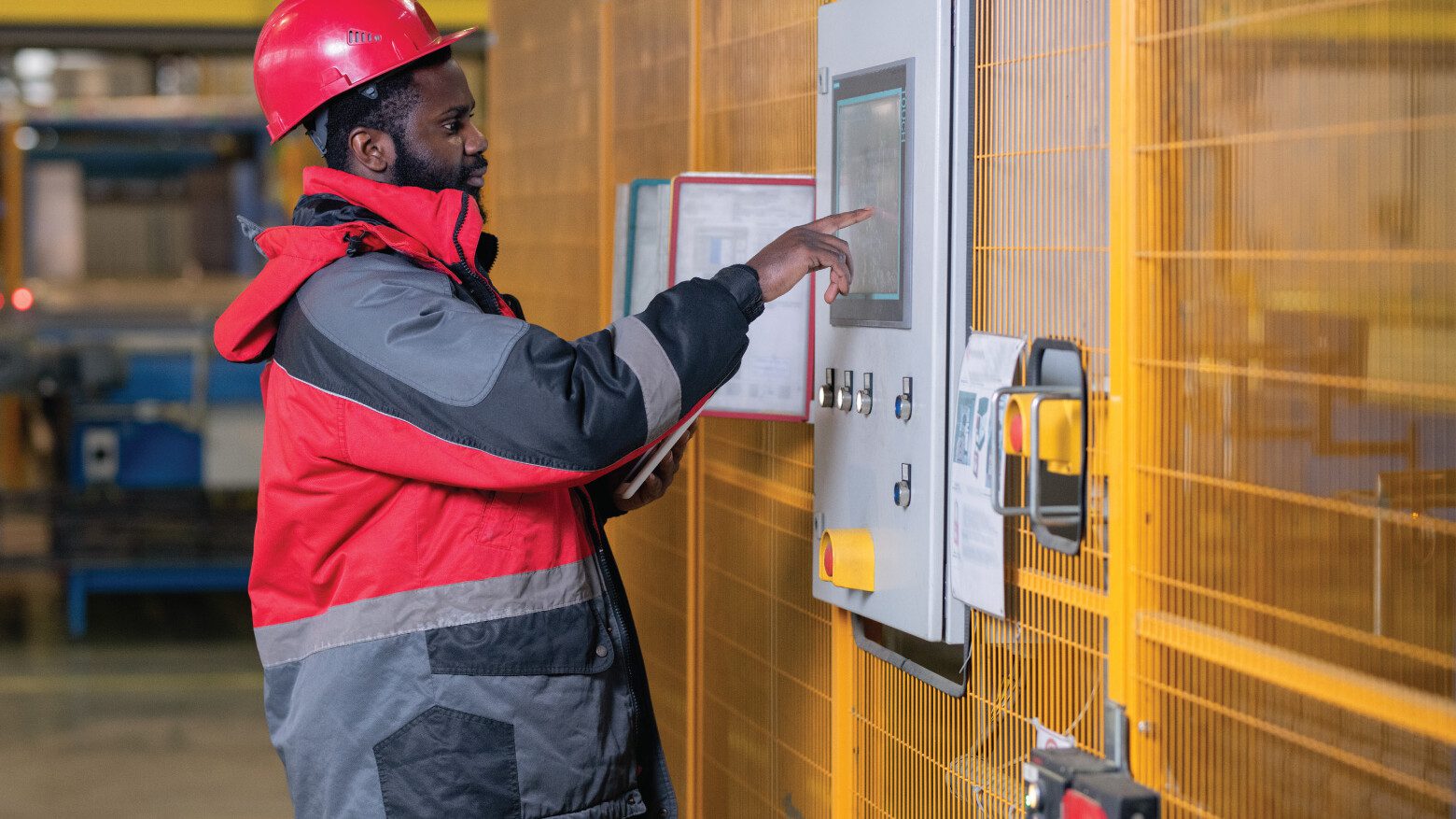 Man in red jacket and hardhat touching a control panel in a warehouse facility